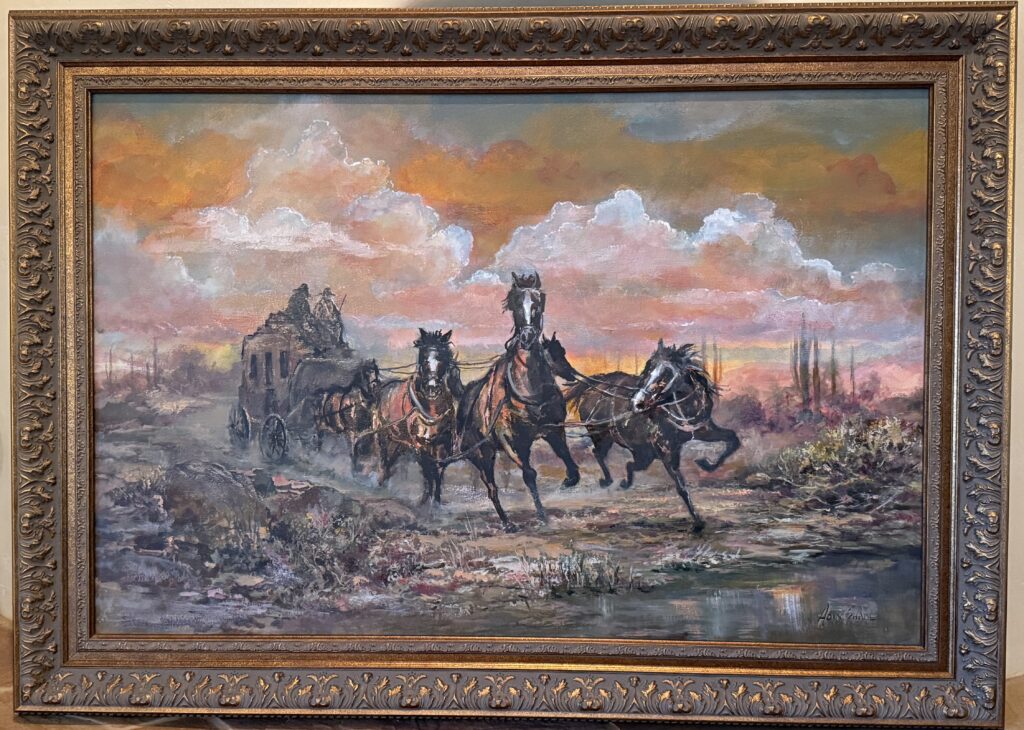 Framed oil painting depicting a team of horses galloping with a stagecoach down a dirt road, set against a desert landscape with saguaro cacti and billowing clouds lit by sunset.