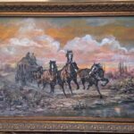 Framed acrylic painting depicting a team of horses galloping with a stagecoach down a dirt road, set against a desert landscape with saguaro cacti and billowing clouds lit by sunset.